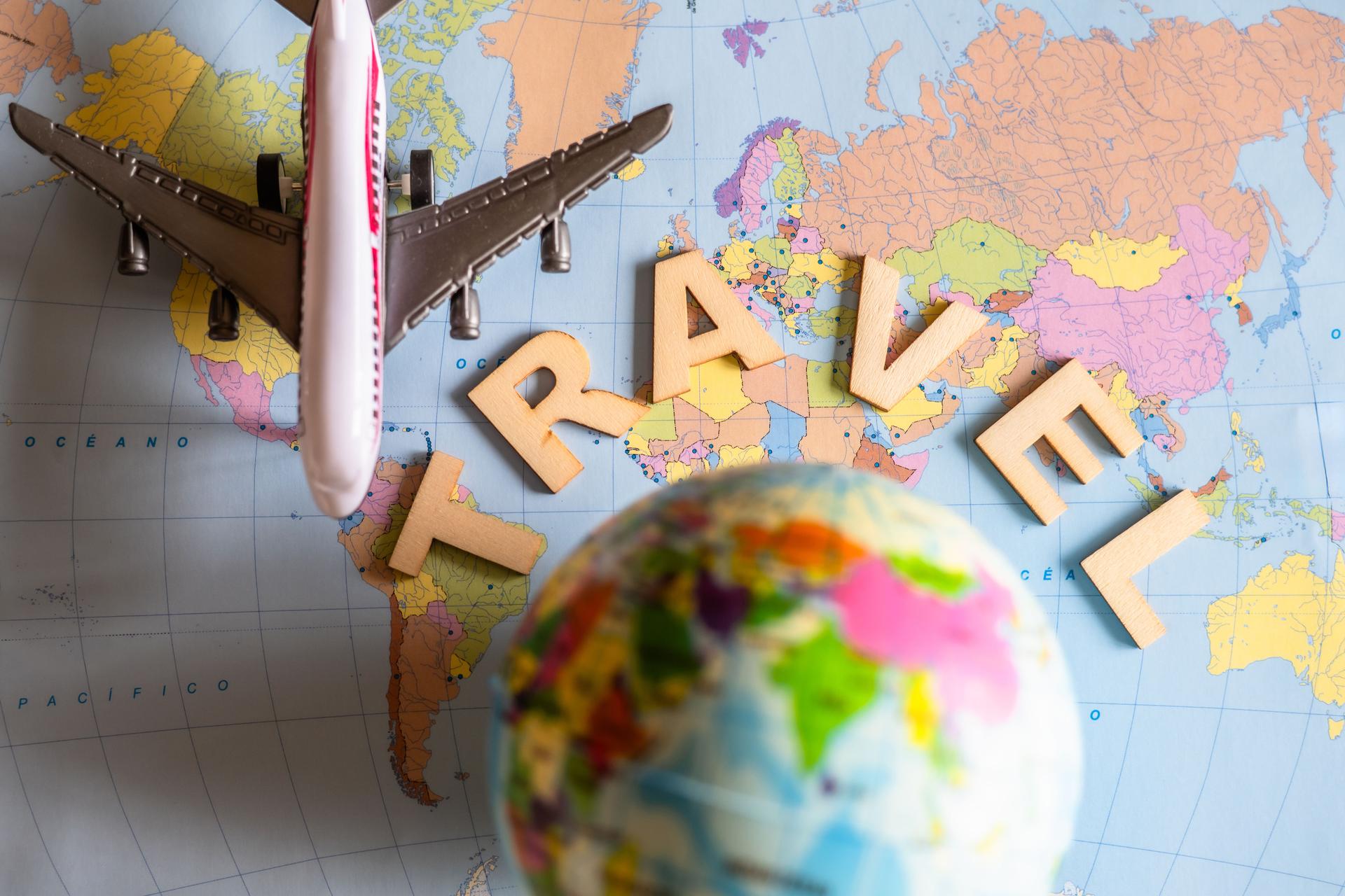 Wooden letters composing the word travel are laying on a colorful world map next to a small toy airplane and a globe, suggesting the planning of a summer vacation trip